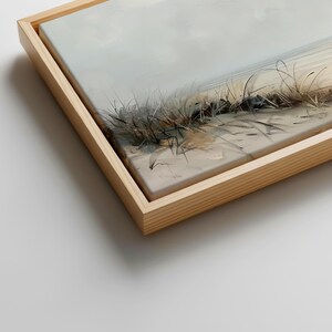 May include: A framed canvas print featuring a coastal landscape with tall, thin dune grasses in shades of brown and grey set against a sandy beach and a soft, muted sky. The artwork is encased in a light-toned, natural wood floating frame.