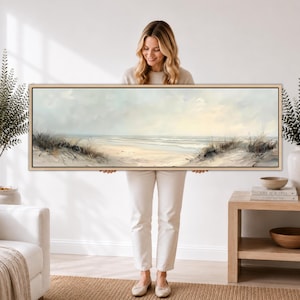 May include: A panoramic landscape painting depicting a serene beach scene with soft sand dunes, gentle ocean waves, and a pale, overcast sky. The artwork is presented in a thin, light-colored wooden frame, showcasing muted tones of beige, cream, and blue.