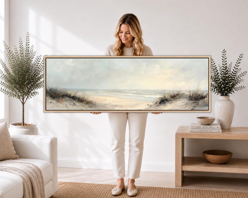 May include: A panoramic landscape painting depicting a serene beach scene with soft sand dunes, gentle ocean waves, and a pale, overcast sky. The artwork is presented in a thin, light-colored wooden frame, showcasing muted tones of beige, cream, and blue.