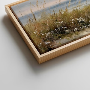 May include: A framed canvas print featuring an impressionistic landscape painting of coastal dunes with tall golden grasses and small white wildflowers under a soft sky, encased in a light-toned wooden frame with a visible grain texture.