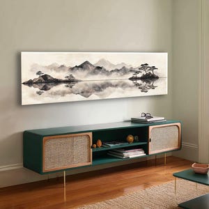 May include: A long, rectangular landscape painting depicts a serene mountain scene in shades of gray and white, with a reflective lake. Below, a dark green cabinet with woven rattan doors and gold legs adds a touch of modern design.