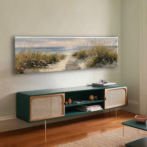 May include: A long, rectangular media console featuring a dark green frame, two side cabinets with light-colored woven cane panels, and an open center shelf. Above the console hangs a wide, horizontal canvas painting of a beach path.