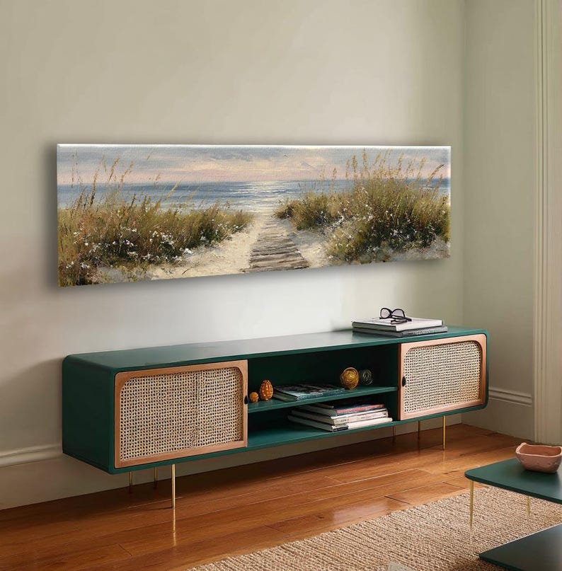 May include: A long, rectangular media console featuring a dark green frame, two side cabinets with light-colored woven cane panels, and an open center shelf. Above the console hangs a wide, horizontal canvas painting of a beach path.