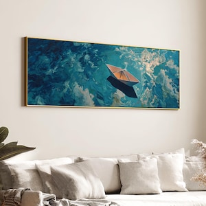 Puede incluir: Una obra de arte enmarcada representa un barco de papel flotando sobre un mar de remolinos azules y blancos. El barco es de color naranja cálido, proyectando una sombra sobre el agua. El marco es dorado y la obra se exhibe sobre un sofá.