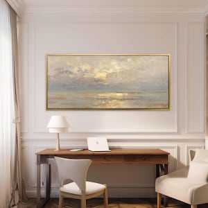 Puede incluir: Pintura abstracta de paisaje con marco dorado. La obra representa un paisaje marino con cielo nublado y luz dorada reflejada en el agua. El cuadro cuelga sobre un escritorio de madera con un portátil, una lámpara y una silla.