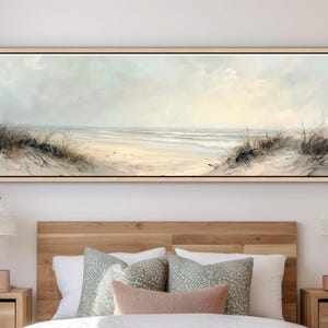 May include: A panoramic landscape painting in a light wood frame depicting a coastal beach scene with sand dunes, tall beach grass, and a calm ocean under a soft, pale sky. The artwork features muted beige, cream, and light blue tones.
