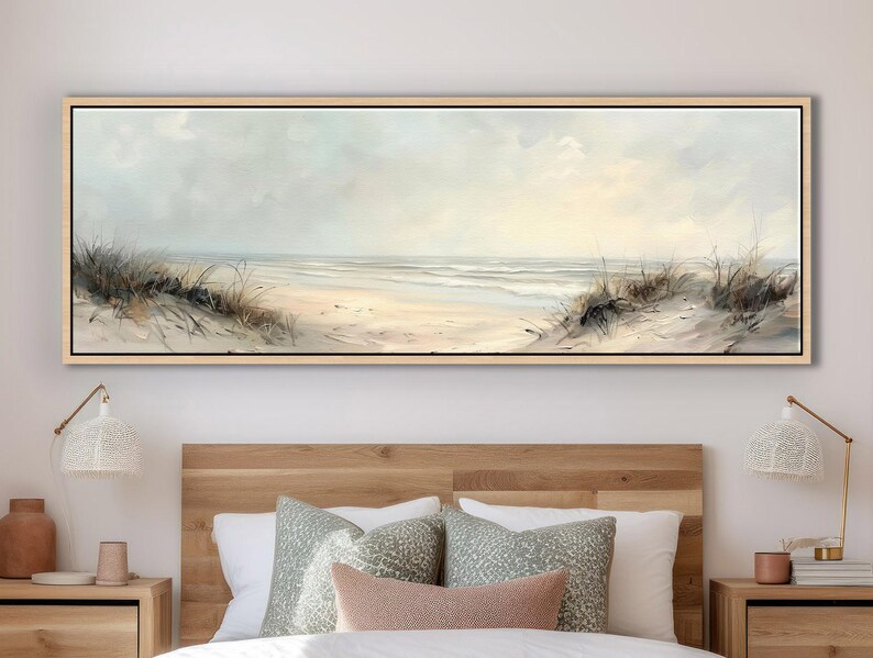 May include: A panoramic landscape painting in a light wood frame depicting a coastal beach scene with sand dunes, tall beach grass, and a calm ocean under a soft, pale sky. The artwork features muted beige, cream, and light blue tones.