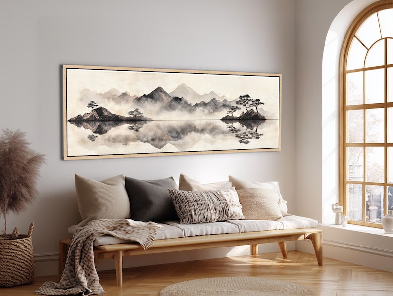 May include: A framed landscape art print depicts a serene mountain scene with a lake. The artwork features a muted color palette of black, gray, and beige, with reflections in the water. The print is displayed above a wooden bench with pillows and a blanket.