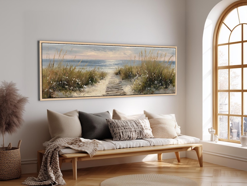 May include: A panoramic landscape painting in a light wood frame depicting a sandy beach path leading toward the ocean. The scene features tall dune grasses, white flowers, and a calm sea under a soft sky, rendered in muted blue, beige, and green tones.
