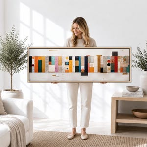 Op de afbeelding: Een lang, rechthoekig ingelijst kunstwerk voor aan de muur met een horizontale opstelling van verticale, veelkleurige houten blokken. De compositie bevat diverse tinten oranje, blauw, zwart, wit en goud tegen een witte achtergrond.