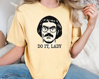 Puede incluir: Camiseta amarillo claro con un gráfico en blanco y negro de un hombre con gafas y bigote. El texto "DO IT, LADY" está impreso debajo. La camiseta es de estilo casual.