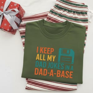 May include: Green crew neck t-shirt with the text "I KEEP ALL MY DAD JOKES IN A DAD-A-BASE" in orange, yellow, teal, and orange. The shirt is paired with striped pyjama bottoms in red, white, and green. A wrapped gift box is in the upper left corner.