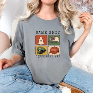 Peut inclure: T-shirt gris avec le texte "SAME SHIT DIFFERENT HAT" et quatre images de chapeaux. Les images incluent un capuchon blanc, un chapeau avec le drapeau confédéré, un chapeau militaire et un chapeau "Make America Great Again".