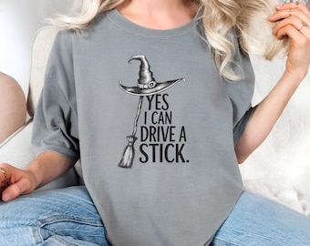 Puede incluir: Camiseta gris con el texto "YES I CAN DRIVE A STICK". El diseño presenta un sombrero de bruja y una escoba. La camiseta es de un estilo cómodo e informal, perfecta para Halloween o un evento temático de brujas.