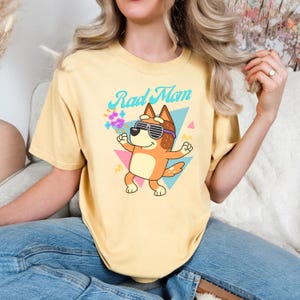 May include: Yellow t-shirt with the text "Rad Mom" and a cartoon dog wearing sunglasses. The dog is orange and brown, with a blue and pink geometric background. The shirt is a light yellow color.