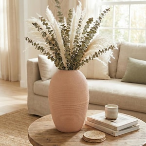 May include: A peach-coloured, textured ceramic vase filled with dried pampas grass and eucalyptus branches. The vase sits on a round, wooden coffee table with books, a candle, and coasters. A neutral-toned sofa is in the background.