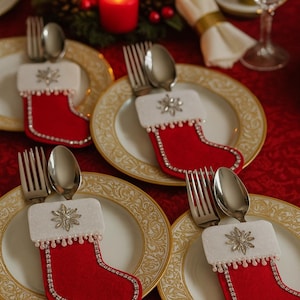 Puede incluir: Una mesa festiva con cuatro platos, cada uno adornado con un portacubiertos en forma de calcetín rojo. La cubertería, incluyendo tenedores y cucharas, descansa sobre los platos. Velas rojas y un centro de mesa decorativo.