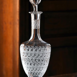 Puede incluir: Una jarra de cristal transparente con un diseño grabado. La jarra tiene un cuello largo y un cuerpo bulboso, con un tapón. El patrón grabado cubre el cuerpo y el cuello, creando un efecto decorativo. Probablemente se utiliza para servir vino u otras bebidas.