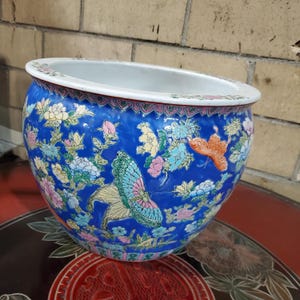 Puede incluir: Una maceta redonda de cerámica azul con un borde ancho. La maceta está decorada con coloridos diseños florales y de mariposas. El borde tiene un borde rosa y blanco. La maceta está sobre una superficie roja.