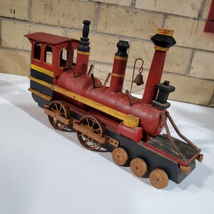 Puede incluir: Modelo de tren de madera pintado de rojo, negro y amarillo. El tren tiene cabina, chimeneas, ruedas y una campana. Un objeto decorativo de estilo vintage.