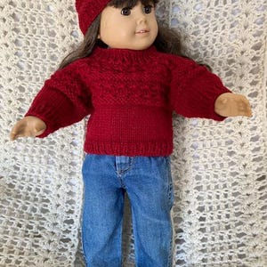 May include: A red knitted sweater and matching hat for a doll. The sweater has a cable knit pattern and the hat is a simple beanie style.