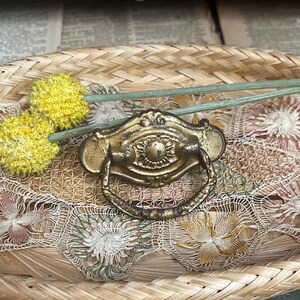 May include: A brass-toned metal drawer pull featuring an ornate, oval-shaped backplate with a central sunburst motif and a swinging, rounded handle. The hardware has a weathered, textured finish and is positioned alongside two yellow flowers.