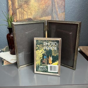 May include: A collection of vintage photo frames in various sizes. The frames are gold-toned with ornate detailing. One frame displays the text "PHOTO FRAME" and the dimensions 3.5 x 5 inches. The frames are arranged on a gray surface.