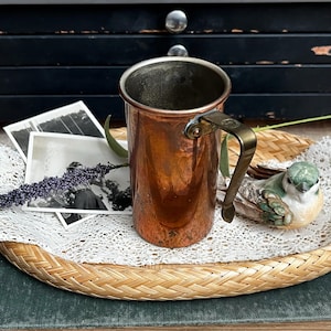 May include: A vintage copper mug with a brass handle. The mug has a shiny, reflective surface and a cylindrical shape. It sits on a woven tray with a lace doily, alongside a small bird figurine and vintage photographs.