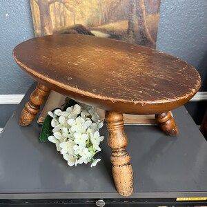 May include: A small, oval-topped wooden stool with three turned legs. The stool is stained a dark brown with visible wear and tear. A bouquet of white flowers rests beneath the stool.