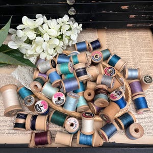 May include: A collection of vintage wooden spools of thread in various colors, including blue, green, red, and white. The spools are arranged in a basket and some are spilling out. The spools are labeled with brand names like "Talon" and "Mercer".