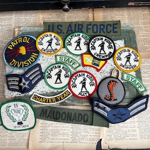 May include: A collection of vintage military patches, including "U.S. AIR FORCE," "Captain Kidd," and various rank insignia. The patches feature embroidered designs in colors like blue, green, and yellow, with text and illustrations.