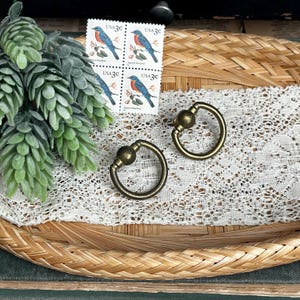 May include: Two antique brass ring pulls with a decorative ball detail, set on a woven tray with a white lace doily. Four vintage 3-cent stamps featuring a bluebird are also present, along with a sprig of green foliage.