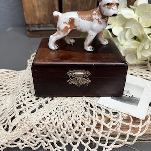 May include: A small, white and brown porcelain dog figurine sits on top of a dark wood box with a metal latch. The box is sitting on a white crocheted doily.