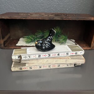 May include: A glossy black ceramic bird figurine with a gold beak rests on a vintage folding ruler. The ruler is white with black markings and measures in inches. The bird is positioned in front of a wooden box.