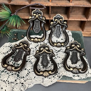 May include: Five ornate, antique-style, gold-toned metal door pulls with a decorative, scrolled design. The pulls have a ring-shaped handle and are arranged on a white crocheted doily.