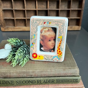 May include: A white ceramic picture frame with a colorful jungle scene and a photo of a young girl. The frame has a pink background with green leaves, a giraffe, a lion, a turtle, and birds.