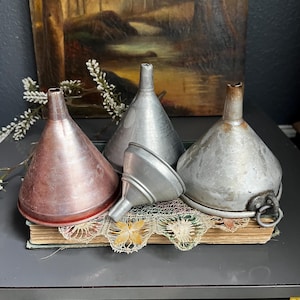 May include: Four antique metal funnels in varying sizes and finishes. The funnels are made of copper and galvanized metal, with a range of colors from reddish-brown to silver. They are arranged on a vintage book with lace trim, suggesting a rustic or vintage theme.