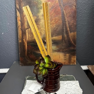 May include: A dark brown ceramic pitcher with a textured, scalloped rim pattern. Inside the pitcher are three yellow wooden folding rulers with black measurement markings and a green and purple artificial succulent plant with thick, fleshy leaves.