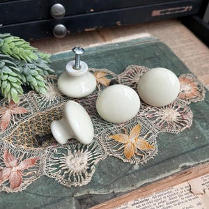 May include: Four off-white ceramic cabinet knobs. Two are spherical, and two have a rounded, flat front with a screw. The knobs are arranged on a vintage lace doily with floral embroidery. A sprig of faux greenery is visible.