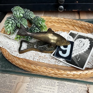 May include: A vintage brass bottle opener shaped like a fish, with a matching brass fish figurine on top. The items rest in a woven basket with a lace doily and a small sprig of greenery. A black and white photo and a number 9 are also in the basket.