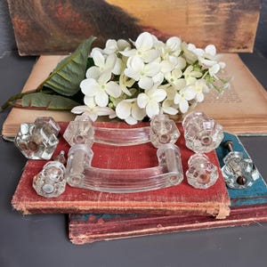 May include: A collection of vintage clear glass cabinet knobs and pulls. The knobs have faceted designs, and the pulls are curved. The items are arranged on a stack of antique books with a red cover, and white flowers.