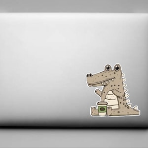 May include: A laptop with a sticker of a cartoon alligator holding a coffee cup. The alligator is light brown with dark brown spots and a white belly. The coffee cup is white with a green coffee bean logo.