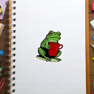 May include: A green frog sticker holding a red mug. The frog is sitting on a green oval, with a white background. The sticker is on a white grid paper notebook, with office supplies in the background.