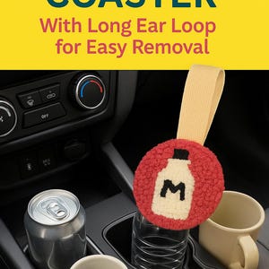 Puede incluir: Posavasos hecho a mano con un diseño rojo texturizado y un gráfico de botella de leche crema. El posavasos tiene un lazo largo beige para una fácil extracción. La imagen también muestra una lata de refresco y una taza de café en el portavasos de un coche.