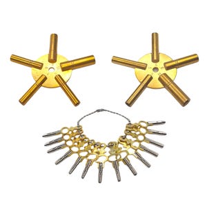 Op de afbeelding: Een verzameling horlogereparatietools. Twee goudkleurige, stervormige tools met genummerde wijzerplaten en elk vijf cilindrische verlengingen. Daaronder een ring van kleine, goudkleurige sleutels met zilveren punten.