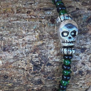 May include: A beaded bracelet with a silver skull charm. The bracelet features alternating black and green beads. The skull charm has eye sockets, a nose cavity, and teeth. The bracelet is displayed on a piece of wood.
