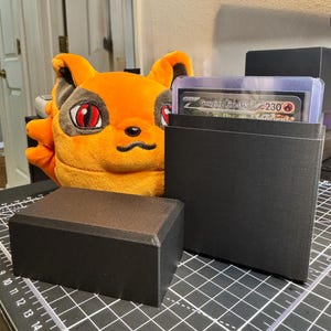 May include: Black card storage boxes with a plush orange fox head in the background. One box holds a card with the text "Gouging Fire 230 Ancient". The boxes are on a black cutting mat.