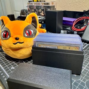 May include: A black card storage box with a lid, holding a stack of trading cards in protective sleeves. An orange plush fox toy with red eyes is in the background. Other items include a black box and a yellow toy car.