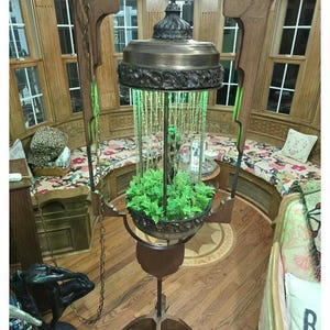 May include: An ornate, antique-style lamp with a bronze-colored top and base. The lamp features a central glass enclosure with green lighting and a plant display. The lamp is suspended from a chain and has a wooden base. The room has wooden paneling and floral patterned cushions.