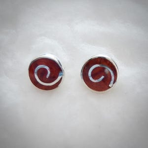 Puede incluir: Un par de pendientes de botón redondos. Cada pendiente presenta un centro rojo oscuro con un diseño de espiral blanco y un borde plateado. Los pendientes se muestran sobre una superficie blanca texturizada.